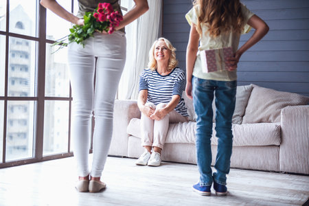 Beautiful senior woman is sitting on couch and smiling while her daughter and granddaughter are holding presents for herの写真素材
