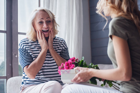 Beautiful senior woman is sitting on couch and smiling while her adult daughter is giving her presentsの写真素材