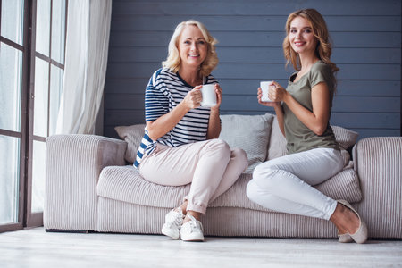 Beautiful senior mom and her adult daughter are holding cups, looking at camera and smiling while sitting on couch at homeの写真素材