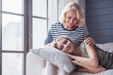 Beautiful senior mom is stroking her adult daughter that lying on her knees, both are smiling while sitting on couch at homeの写真素材