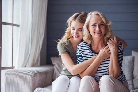 Beautiful senior mom and her adult daughter are hugging and smiling while sitting on couch at homeの写真素材