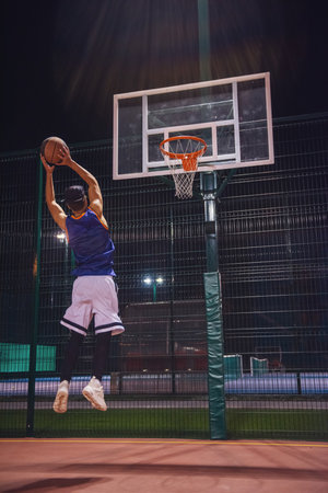 Full length portrait of stylish young basketball player in cap jumping and shooting a ball through the hoop while playing outdoors at nightの写真素材