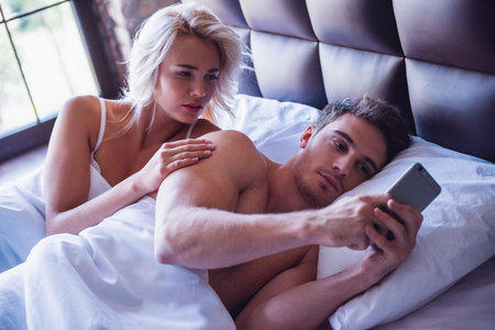 Beautiful young couple is lying in bed, guy is using a smart phone, girl is looking with reproachの写真素材