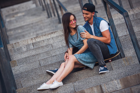 Stylish young couple is hugging, talking and smiling while sitting on the skateboard on stairsの写真素材