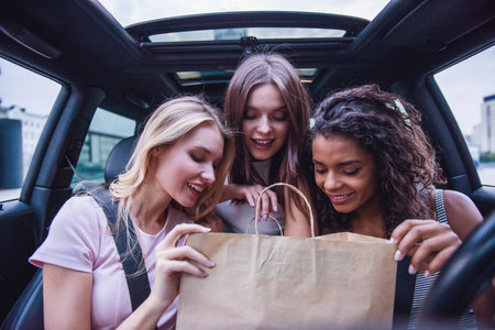 Cheerful beautiful girls sitting in the car, looking at shopping in a paper bag
