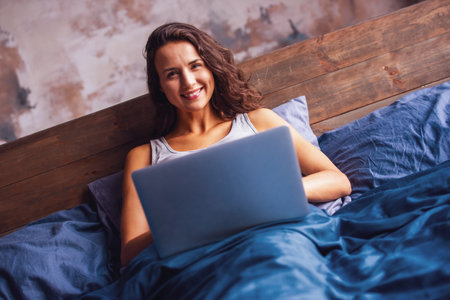 Beautiful young woman is looking at camera and smiling while working with a laptop in bed in the morningの写真素材