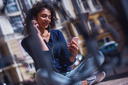 Attractive girl in casual clothes and earphones is listening to music using a smart phone and...
