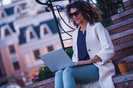 Attractive young woman in white coat and sun glasses is using a laptop and smiling while resting in the cityの写真素材