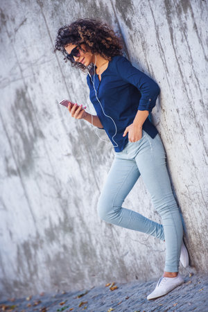 Attractive girl in casual clothes, glasses and earphones is listening to music using a phone and smiling while standing outdoors near the wallの写真素材