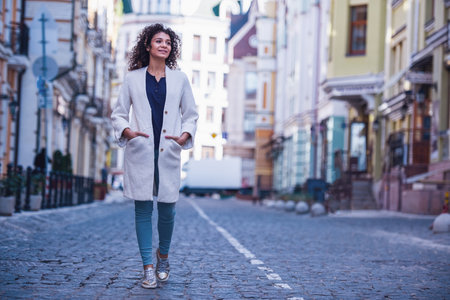 Attractive young woman in white coat is smiling while walking in cityの写真素材