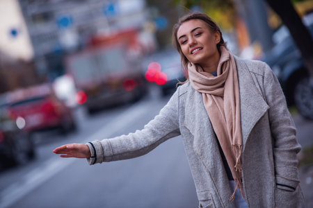 Beautiful girl in coat is smiling and lifting her hands while catching a taxi in the cityの写真素材