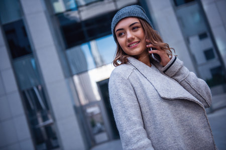 Beautiful girl in coat and cap is talking on the mobile phone and smiling while walking in the cityの写真素材