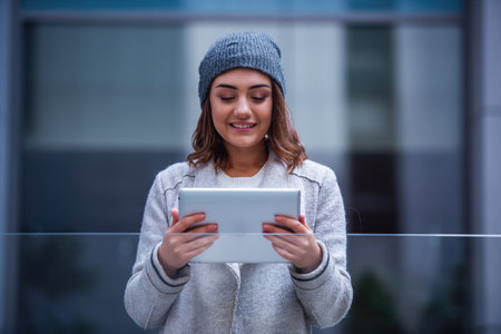 Beautiful girl in coat and cap is using a digital tablet and smiling while resting outdoorsの写真素材