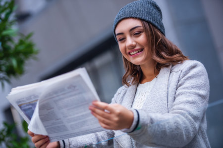 Beautiful girl in coat and cap is reading a newspaper and smiling while resting outdoorsの写真素材