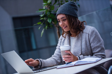 Beautiful girl in coat and cap is drinking coffee and smiling while working with a laptop in a cafe outdoorsの写真素材