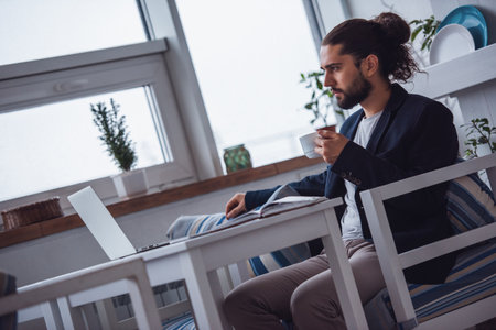 Handsome young businessman with long dark hair is drinking coffee while working with a laptop in a cafeの写真素材
