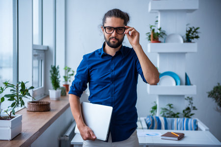 Stylish young businessman with long dark hair is holding a laptop and looking at camera while working in the cafeの写真素材