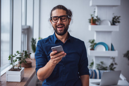 Stylish young businessman with long dark hair is using a smartphone and laughing while working in the cafeの写真素材