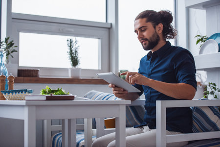 Handsome young man with long dark hair is using a digital tablet and smiling while eating in the cafeの写真素材