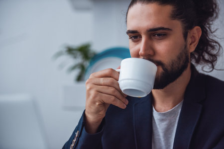 Handsome young businessman with long dark hair is drinking coffeeの写真素材