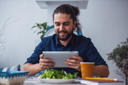Handsome young man with long dark hair is using a digital tablet and smiling while eating in the cafeの写真素材