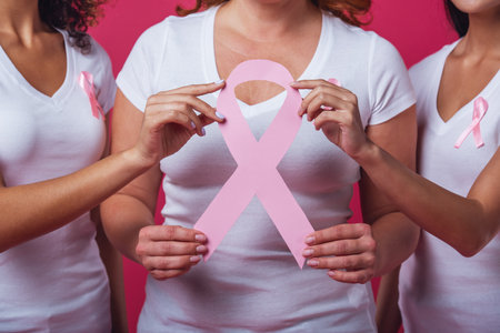 Women against breast cancer. Beautiful women with pink ribbons on their chests holding a pink paper ribbon and smiling, cropped, on red backgroundの写真素材