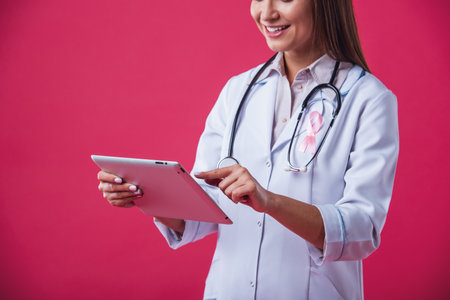 Women against breast cancer. Cropped image of beautiful female doctor with a pink ribbon on her chest is using a tablet and smiling, on red backgroundの写真素材
