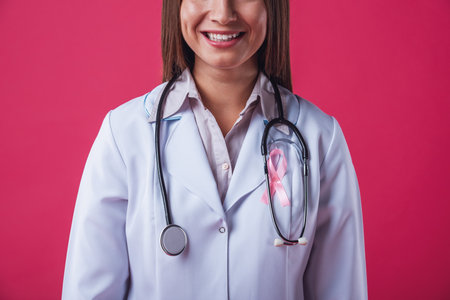 Women against breast cancer. Cropped image of beautiful female doctor with a pink ribbon on her chest smiling, on red backgroundの写真素材