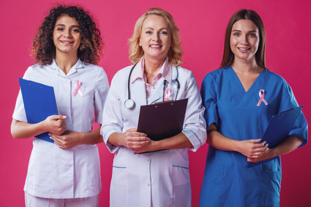 Women against breast cancer. Beautiful young female doctors with pink ribbons on their chests are holding folders, looking at camera and smiling, on red backgroundの写真素材