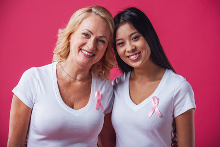 Women against breast cancer. Beautiful mature and Asian women with pink ribbons on their chests are looking at camera and smiling, on red backgroundの写真素材
