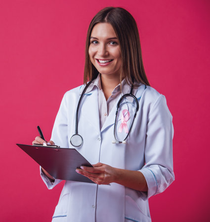 Women against breast cancer. Beautiful female doctor with a pink ribbon on her chest is holding a folder, looking at camera and smiling, on red backgroundの写真素材