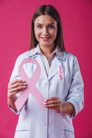 Women against breast cancer. Beautiful female doctor is holding a pink ribbon, looking at camera and smiling, on red backgroundの写真素材