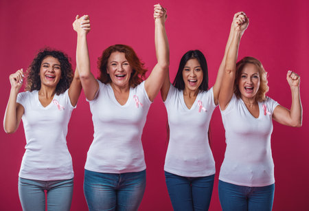 Women against breast cancer. Beautiful women of different ages and nationalities with pink ribbons on their chests are raising hands and smiling, on red backgroundの写真素材