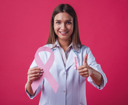 Women against breast cancer. Beautiful female doctor is holding a pink ribbon, showing Ok sign and smiling, on red backgroundの写真素材