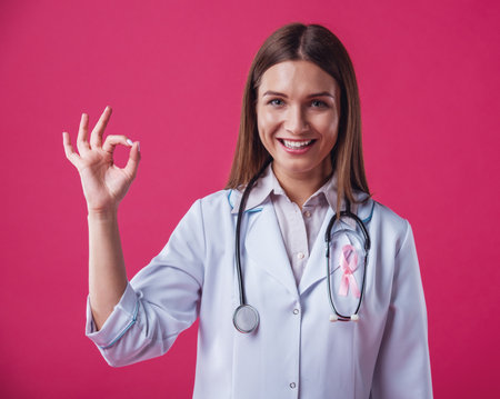 Women against breast cancer. Beautiful female doctor with a pink ribbon on her chest is showing Ok sign, looking at camera and smiling, on red backgroundの写真素材