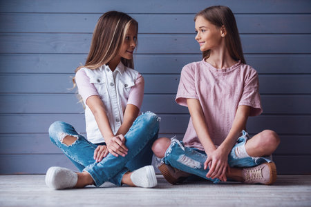 Two attractive teenage girls in casual clothes looking at each other and smiling while sitting on the floor against a gray wall backgroundの写真素材