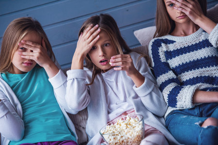 Three attractive teenage girls in casual clothes are eating popcorn and covering their faces while watching TV on couch at homeの写真素材