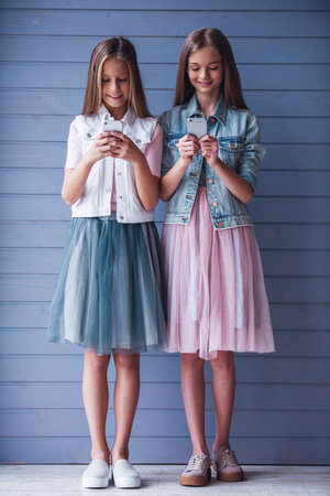 Full length portrait of two attractive teenage girls in dresses using smartphones and smiling, on gray wall backgroundの写真素材
