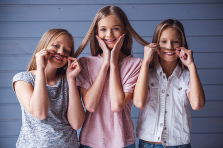 Three attractive teenage girls in casual clothes are looking at camera and smiling, two are making mustaches of their friend's hair, on gray wall backgroundの写真素材