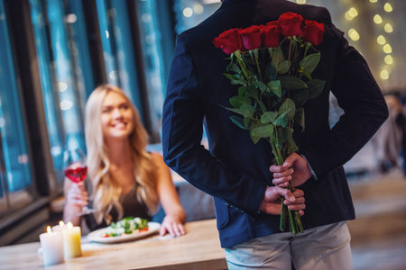 Beautiful girl is sitting at the table in restaurant and smiling while a man is holding roses behind his backの写真素材
