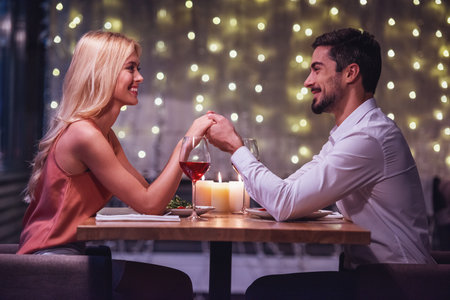Beautiful young couple is looking at each other, holding hands and smiling during their date in a restaurantの写真素材