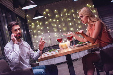 Beautiful young woman is annoyed while her boyfriend is talking on the mobile phone during their date in a restaurantの写真素材