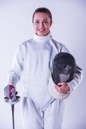 Beautiful female fencer in protective clothing is holding a mask and a weapon, looking at camera and smiling, on gray backgroundの写真素材