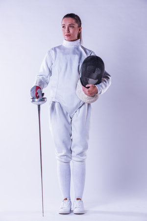 Beautiful female fencer in protective clothing is holding a mask and a weapon, on gray background, full-lengthの写真素材