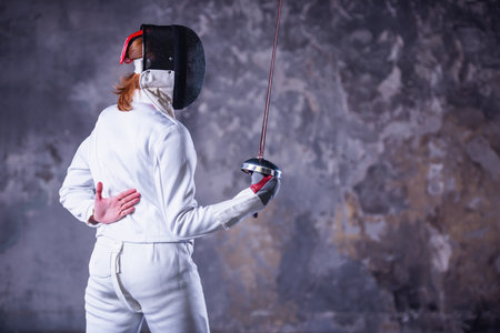 Female fencer in protective clothing is using weapon while practicing on dark gray backgroundの写真素材