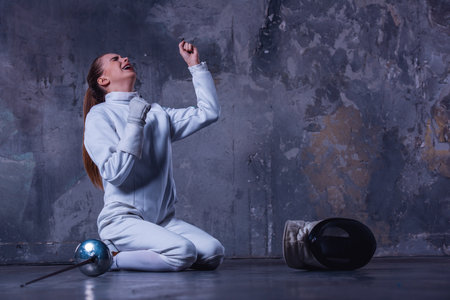 Attractive female fencer in protective clothing is sitting on her knees and screaming with happiness, on dark gray backgroundの写真素材
