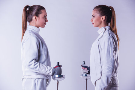 Beautiful female fencers in protective clothing are holding weapon and looking at each other, on gray background, side viewの写真素材