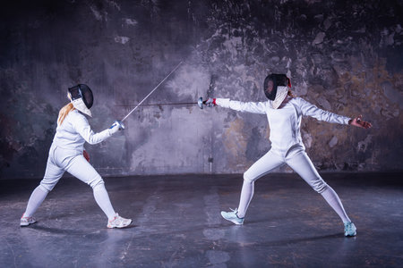 Female fencers in protective clothing are fighting on dark gray background, full-lengthの写真素材