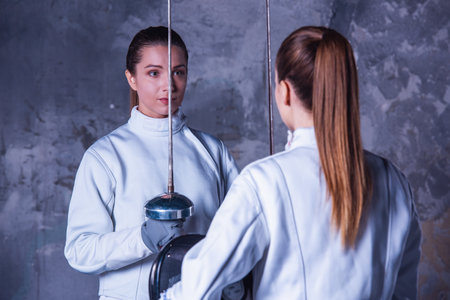 Beautiful female fencers in protective clothing are holding masks and weapon and looking at each other, on dark gray backgroundの写真素材