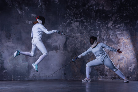 Female fencers in protective clothing are fighting on dark gray background, full-lengthの写真素材